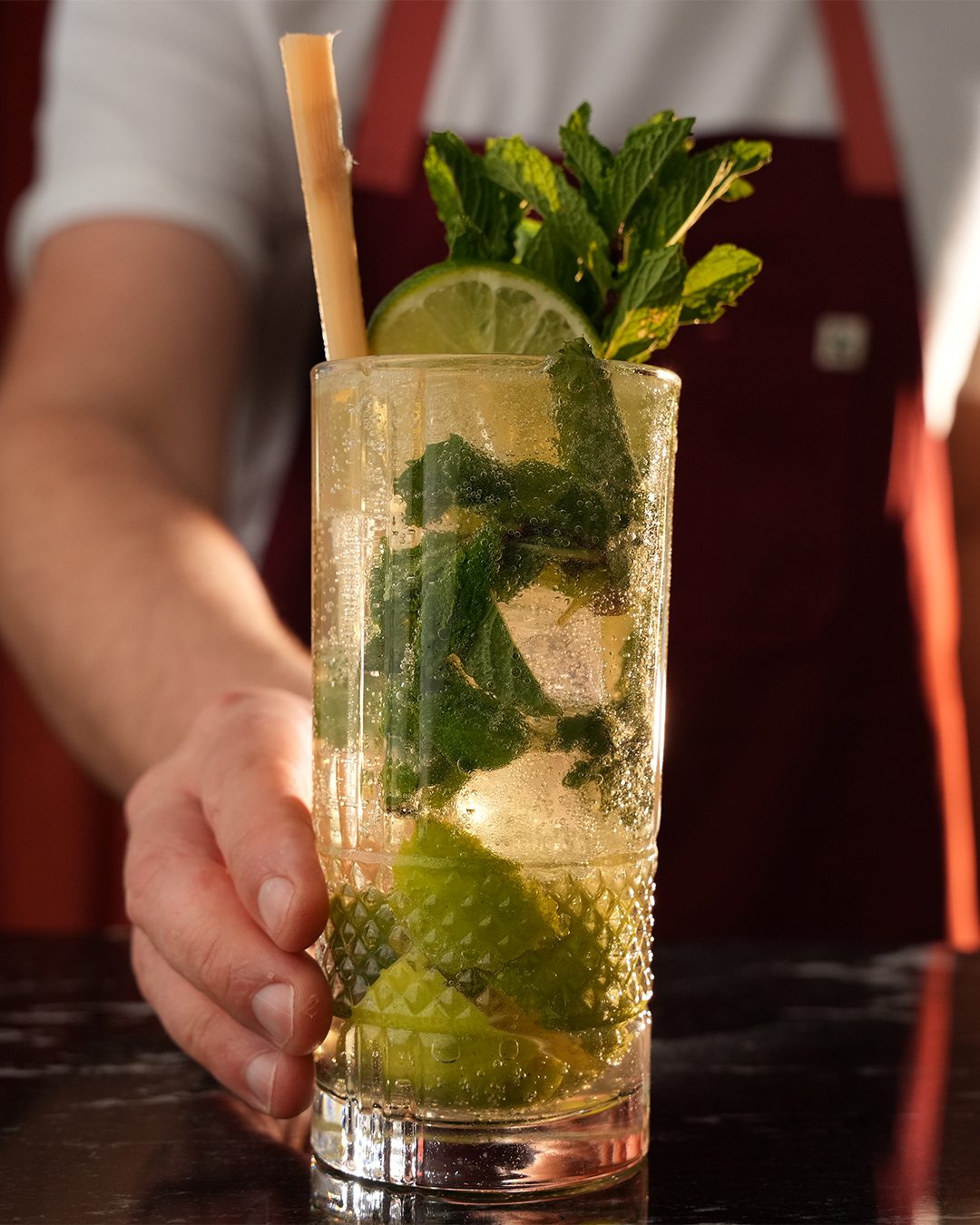 A bartender holds out a Mojito Clásico, a tall glass packed with mint and lime, against Bar Victor's red curtain