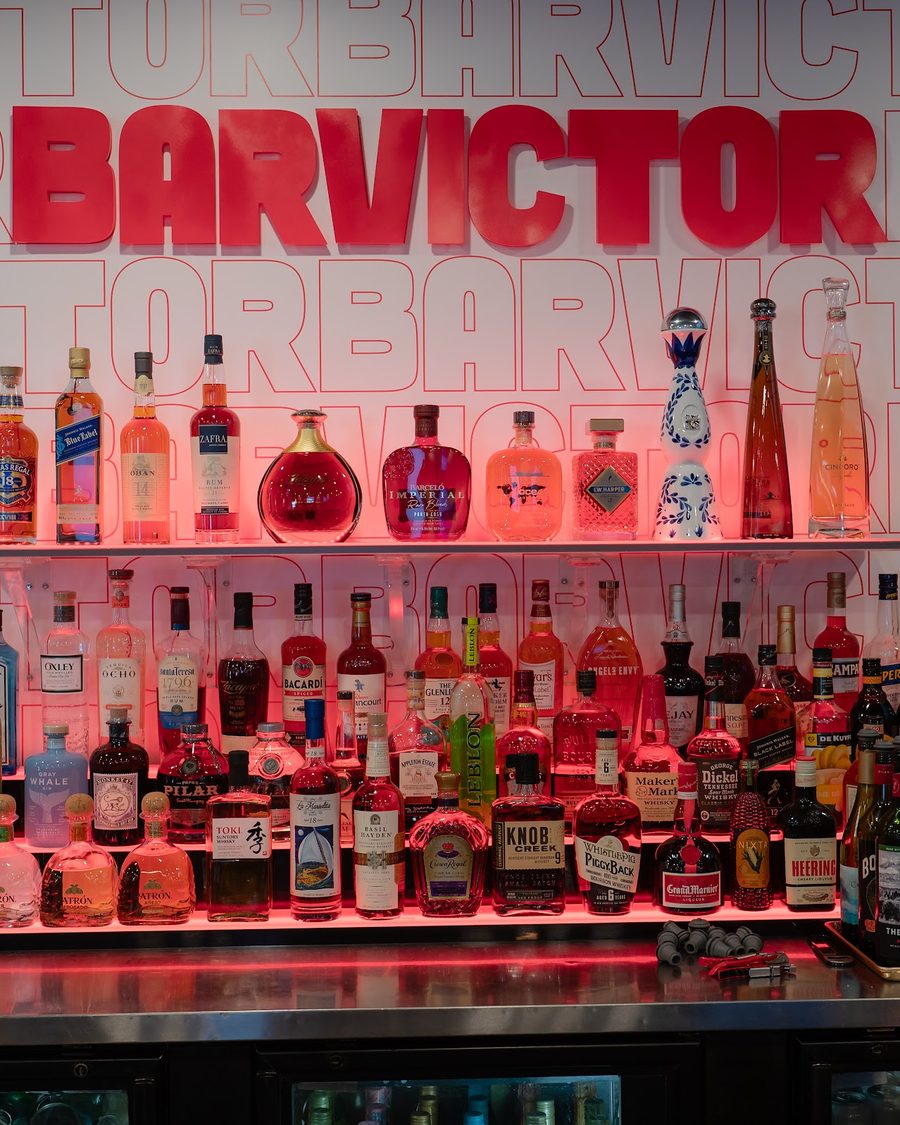 The lit back bar at Bar Victor with rows of bottles under the Bar Victor sign