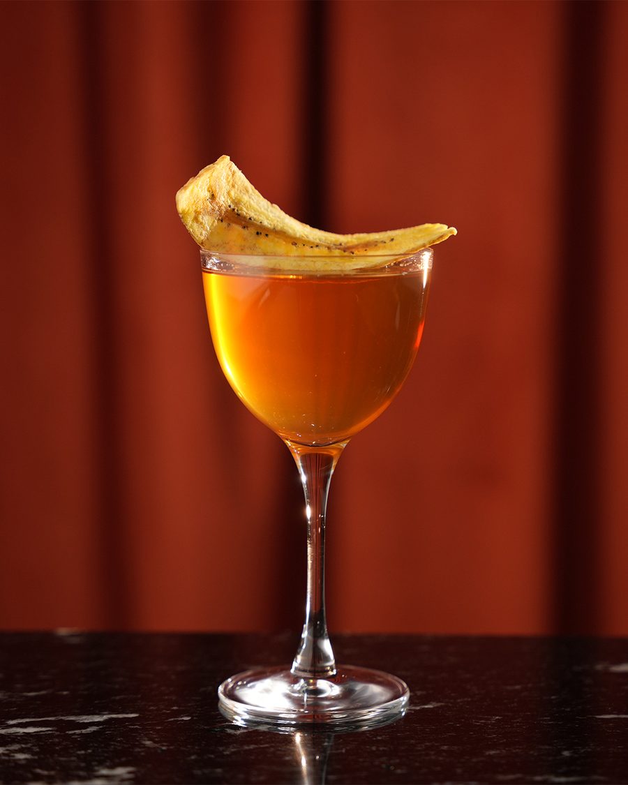 An amber coupe cocktail garnished with a plantain chip