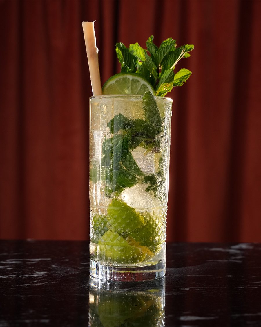 A Mojito Clásico in a tall glass with fresh mint and lime, held out by a bartender